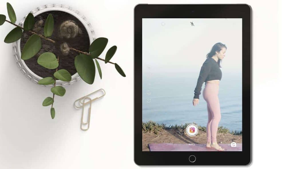 Flat lay of tablet showcasing an Instagram Reel of a woman by the sea. To the left-hand side, a plant and paperclips on a white background. Instagram search and explore